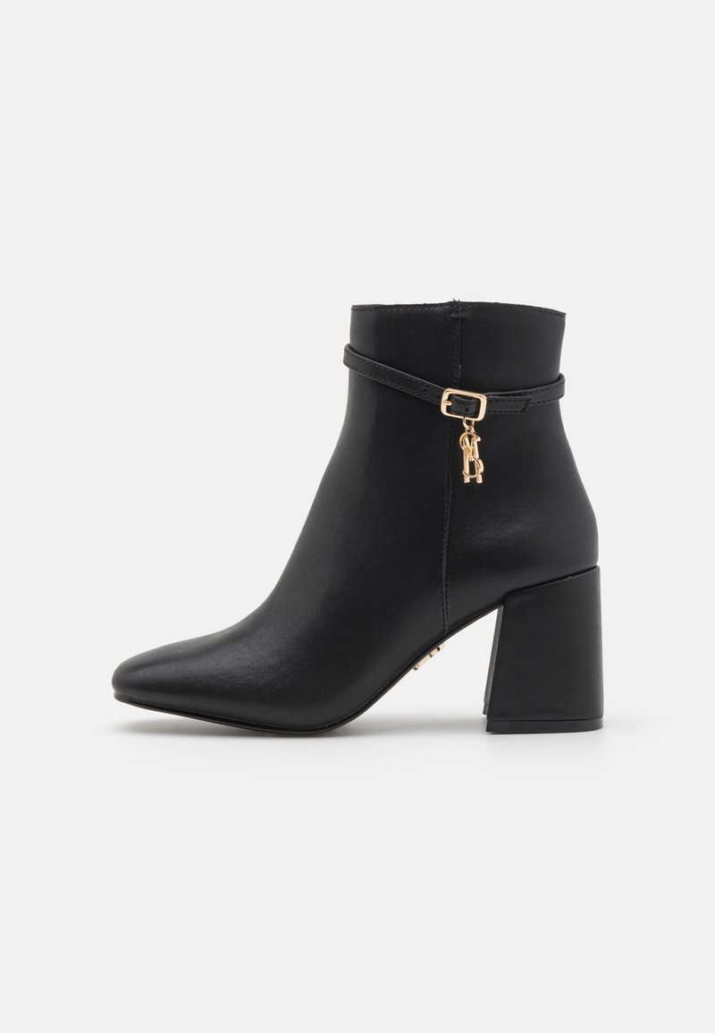Bottine noire en simili cuir lisse, présentant un bout pointu, un talon bloc et une sangle décorative dorée avec un charm.
