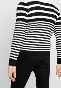 Pull en tricot noir et blanc rayé avec des poignets et un ourlet côtelés, associé à un jean noir avec poches arrière. Design décontracté.