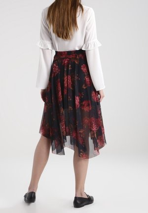 Femme debout, de dos, portant un chemisier blanc et une jupe asymétrique noire à imprimé floral rouge, avec des ballerines noires.