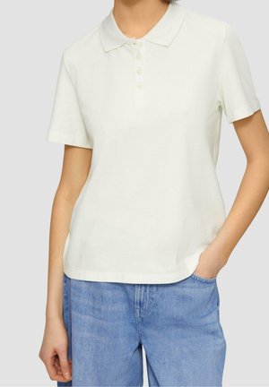 Personne portant un polo blanc uni à manches courtes et un jean bleu clair, une main dans la poche, sur un fond neutre.