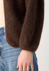 Brauner, flauschiger Pullover mit rundem, puffigem Ärmel-Design, kombiniert mit hellblauen Jeans. Weiche Textur und reichhaltige Farbe.