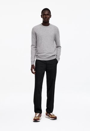 Homme debout portant un pull léger gris clair à manches longues, un pantalon noir et des baskets marron avec des lacets blancs sur un fond blanc uni.