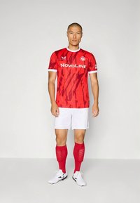 Rød fotballtrøye med et geometrisk mønster, hvite detaljer og logo. Kombinert med hvite shorts og røde knelange sokker. Svarte fotballsko.