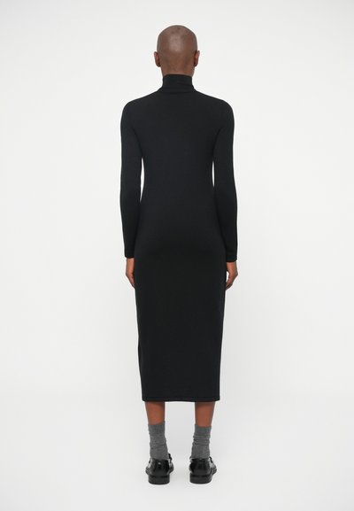 Melnā, gara piedurkne, turtlenecka kleita ar pieguļošu siluetu. Nav redzamu rakstu. Valkāta ar pelēkām zeķēm un melnām kurpēm. Gluda auduma.