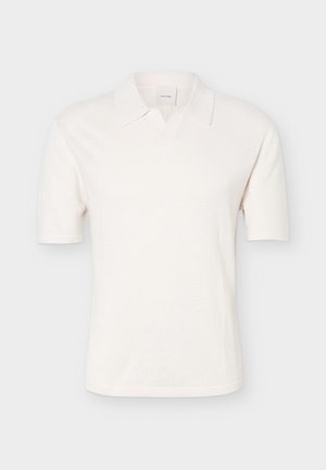 Fehér, rövid ujjú teniszpóló kötött mintával és nyitott gallérral, egyszerű világosszürke háttéren bemutatva.