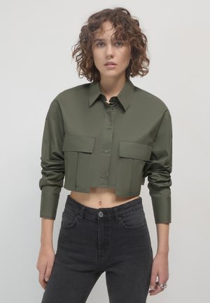 Cropped olive green shirt with button closure and two front pockets, paired with dark gray denim jeans. Smooth fabric and tailored fit.