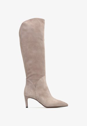 Beżowy zamszowy botek do kolan z ostrym noskiem, smukłą sylwetką i umiarkowanym obcasem. Posiada przeszywane detale oraz gładką teksturę.