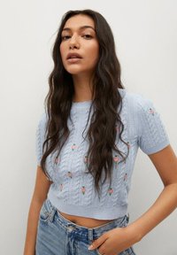 Jasnoniebieski, krótki sweter crop top z krótkim rękawem, o fakturze warkoczowego splotu i wyszywanymi różowymi kwiatami. Posiada karbowany dekolt w kształcie falbanki oraz ściągacz na dole.