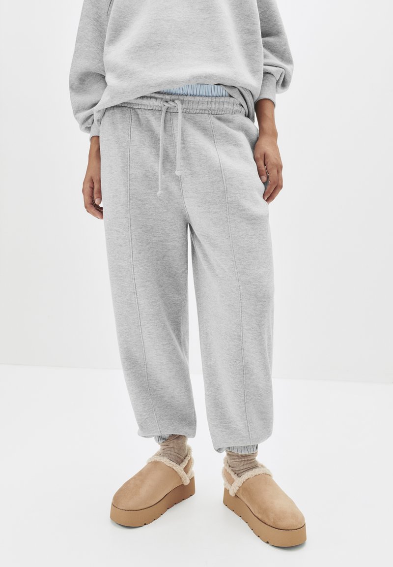 Graue Jogginghosen mit elastischem Bund und Bündchen, die eine weiche Textur und gestickte Details aufweisen, kombiniert mit beigen Clogs gefüttert mit Fell.
