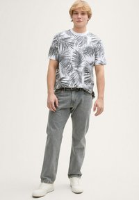 Camiseta blanca con estampado de hojas de palma negras, combinada con jeans de mezclilla gris claro y zapatillas blancas. Diseño limpio, corte casual y detalles mínimos.