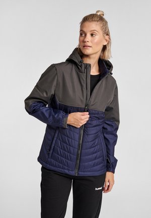 Kvinde med blond hår i en halv knold iført en tofarvet sort og navy hættetrøje, der lynes op, stående mod en ensartet lys baggrund.