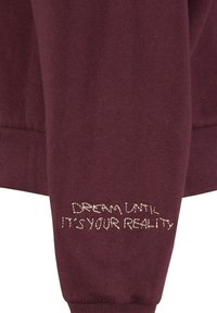 Bordeaux sweatshirt met geribbelde manchetten, met de tekst "DROOM TOT HET JE REALITEIT IS" in lichtbeige geborduurd op de mouw. Zachte textuur.