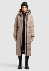 Hosszú bézs színű steppelt dzseki kapucnival, oldalsó cipzárakkal és nagy párnázott panelek.