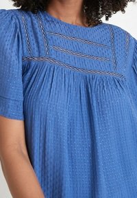 Modrý strukturovaný top s krátkými rukávy, jemnými krajkovými detaily a nařaseným designem. Měkká tkanina s jemným puntíkovaným vzorem.