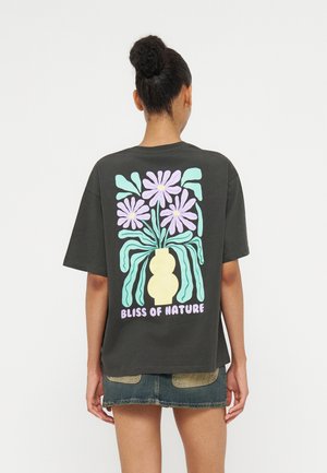 Femme avec les cheveux en chignon porte un T-shirt noir avec un motif coloré de vase à fleurs et le texte "Bliss of Nature" dans le dos, associé à un short en jean.