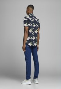 Camisa de flores de manga corta en azul marino con patrones en rosa y verde, combinada con jeans azules y zapatillas blancas, vista desde atrás.