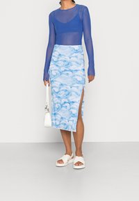 Sininen läpinäkyvä pitkähihainen paita, vaaleansininen hame aalto-kuviolla, sivuhalkio, valkoiset sandaalit ja pieni valkoinen käsilaukku.