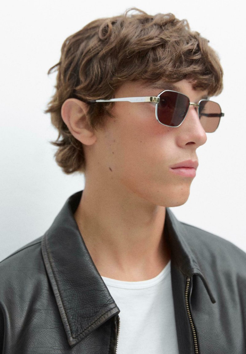 Metaliczne prostokątne okulary przeciwsłoneczne z ciemnymi soczewkami, cienką srebrną ramką i subtelnymi złotymi akcentami, zestawione z czarną skórzaną kurtką i białą koszulką.