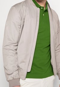 Homme portant une veste beige zippée sur un polo vert et un pantalon clair sur un fond uni.