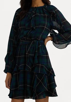 Zelené kostkované šaty s volánkovým designem, dlouhými rukávy a páskem v pase. Vyrobené z lehké látky s vrstvenou sukní a zdůrazněnými švy.