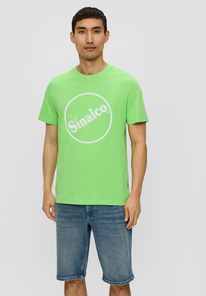 Grünes Baumwoll-T-Shirt mit weißem, rundem "Sinalco"-Logo. Rundhalsausschnitt, kurze Ärmel, kombiniert mit blauen Denim-Shorts, lässige Passform.