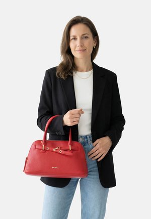 Donna con blazer nero e top bianco tiene una grande borsa in pelle rossa con dettagli dorati su uno sfondo semplice.