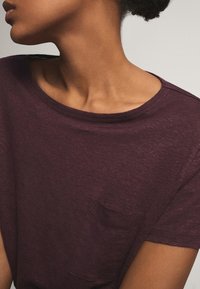 Burgundy T-paita, jossa on pyöreä pääntie ja lyhyet hihat. T-paidassa on pieni rintatasku ja se on valmistettu teksturoidusta, pehmeästä kankaasta.