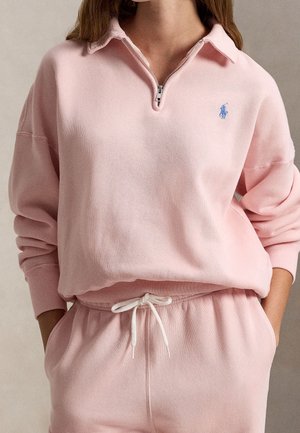 Nainen yllään vaaleanpunainen vetoketjukauluksinen collegepaita, jossa on sininen logo, ja yhteensopivat verkkarit valkoisella kiristysnauhalla, kädet taskuissa.