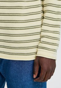 Striped knit sweater in light yellow with green and white lines, featuring a ribbed cuff. Worn with denim blue jeans.