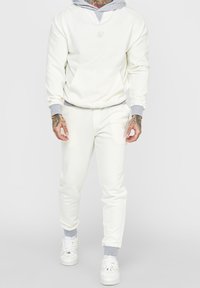 Vit sweatshirt med grå huva och muddar, med en framficka och subtil logotyp. Matchas med vita byxor och vita sneakers.