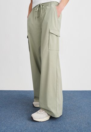 Inimene, kes kannab helerohelisi laia säärega lahtisi pükse nöörvöökohaga ja beeže tenniseid, seisab sinisel vaibal.