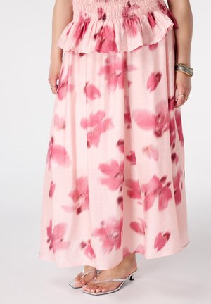 Vrouw draagt een lange roze rok met donkerroze bloemmotieven en volantdetails, gecombineerd met zilveren sandalen met hak en armbanden aan één pols.