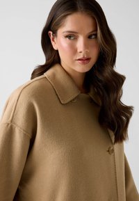 Cappotto beige in misto lana con colletto strutturato, vestibilità oversize e chiusura frontale con bottoni. Texture morbida e dettagli di cuciture minime visibili.