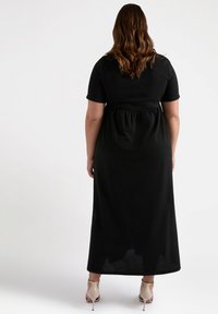 Black maxi dress with short sleeves, gathered waist, flowing skirt, and smooth texture. Worn with beige high-heeled shoes.