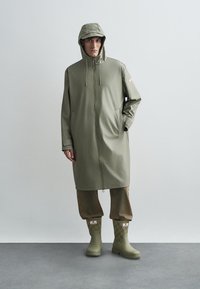 Olivnozeleni vodoodporni dežnik s kapuco, veliki sprednji žepi in zadrgo po dolžini, nošen preko ohlapnih bež hlač in vzorčastih zelenih čevljev.