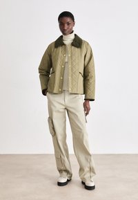 Quilted olive green jacket with a dark collar, gold buttons, and side pockets, paired with beige cargo pants featuring a zippered pocket.