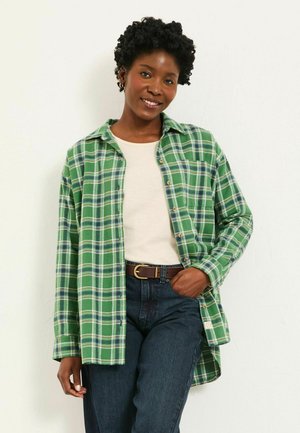 Green plaid shirt with white and navy accents, button-down front, long sleeves, worn over a light-colored top and dark jeans.