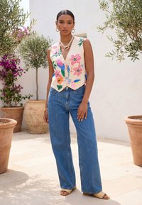 Vrouw staat buiten en draagt een mouwloos vest met bloemenborduursel, een blauwe spijkerbroek, gouden sieraden en gevlochten sandalen, omringd door potplanten.