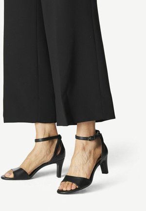 Mujer con sandalias negras de tacón alto con tiras en el tobillo, dedos abiertos y pantalones negros de pernera ancha recortados por encima de los tobillos.