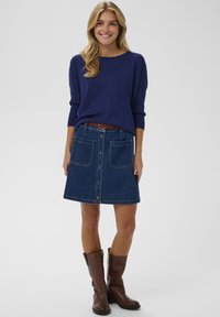 Pull en maille bleu marine associé à une jupe trapèze en denim foncé avec fermeture boutonnée sur le devant et deux grandes poches plaquées. Ceinture et bottes en cuir marron.