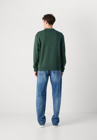 Pulover verde cu guler rotund, croială relaxată și manșete cordonate, purtat cu blugi deschiși la culoare, cu picior drept și adidași albi.
