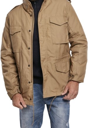 Mężczyzna w beżowej, zapinanej na guziki kurtce z wieloma kieszeniami z patkami, na czarnym swetrze i w wyblakłych niebieskich dżinsach, trzymający krawędzie kurtki.