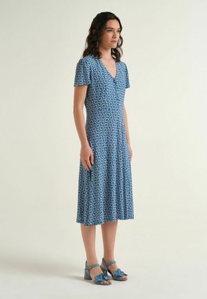 Donna in piedi che indossa un vestito blu fantasia al ginocchio con maniche corte e sandali blu con tacco con dettagli floreali.