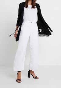 Vita vida-byxor med hög midja, tillsammans med en grå t-shirt och en svart cardigan. Inkluderar svarta klacksandaler.