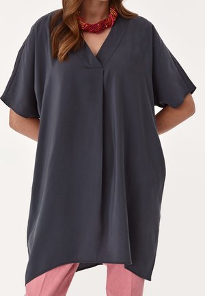 Femme portant une tunique ample gris foncé à col en V avec manches courtes et ourlet asymétrique, sur un pantalon rose, accessoirisée d'un collier de perles rouges.