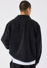 Chaqueta de denim negra con un ajuste relajado, que cuenta con un cuello con solapa, costuras de doble puntada y un dobladillo ligeramente deshilachado en la parte posterior.