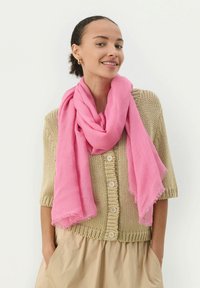Écharpe rose en tissu léger, drapée de manière décontractée autour du cou, avec des bords effilochés, assortie à un cardigan beige en tricot.