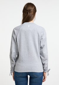 Szürke pulóver hosszú raglan ujjakkal, illeszkedő mandzsettákkal húzózsinórral és gomb részlettel a hát nyaknál. Puha, texturált pamut keverék.