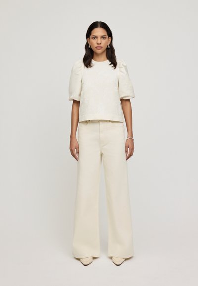 Top con maniche a sbuffo in crema con motivo testurizzato, abbinato a pantaloni larghi in crema e tacchi a punta beige. Design semplice e minimalista.