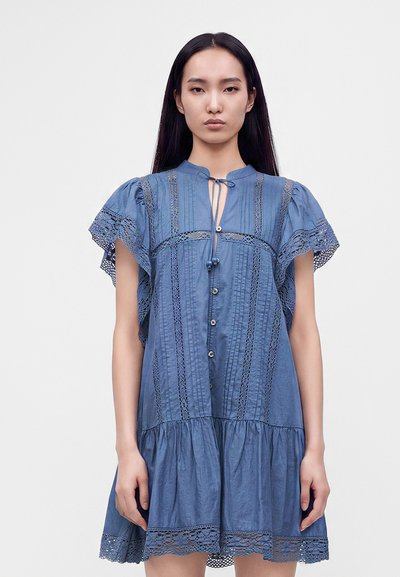 Femme aux longs cheveux noirs portant une robe bleue à manches courtes avec des détails en dentelle et des boutons, debout devant un fond clair uni.